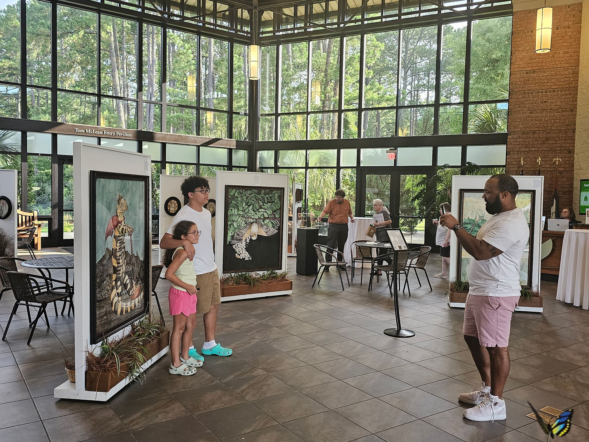 A wide, horizontal photo of an art exhibition inside a building with large windows and a high ceiling. Several framed paintings are on display on white stands. In the foreground, a man in a white t-shirt and pink shorts takes a photo of a painting with his phone. Next to him, a young girl and a teenage boy stand looking at a painting. Other people are milling about in the background.