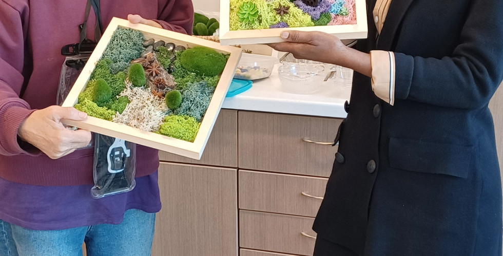 A vertical full-body shot of two women standing and smiling while holding framed pieces of moss art. The woman on the left, with short gray hair, wears a purple hoodie and blue jeans, holding a square frame with various shades of green moss and small faux plants. The woman on the right, with dark hair, wears a black blazer over a light-colored shirt and black pants, holding a similar frame with green and white moss, pinecones, and colorful decorative berries. They are standing in a room with light-colored walls and a cabinet in the background.