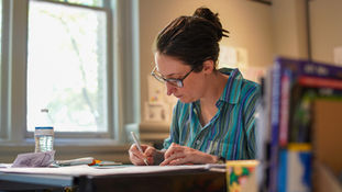 A low-angle, side profile of an artist with their hair in a bun, diligently sketching or inking a project at a desk. The background shows a soft, bright window and various art books.