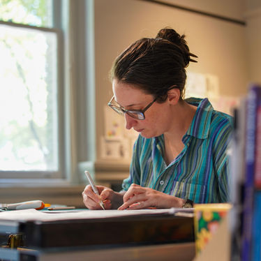 A low-angle, side profile of an artist with their hair in a bun, diligently sketching or inking a project at a desk. The background shows a soft, bright window and various art books.