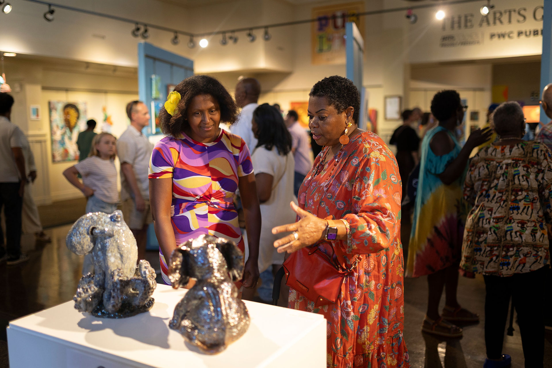  Two women are looking at two ceramic sculptures of dogs on a pedestal in an art gallery. The woman on the left is wearing a colorful patterned dress and has a yellow flower in her hair, while the woman on the right is gesturing as she speaks, wearing a red and orange patterned dress. Other people are in the background.