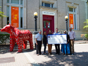 Six adults pose with a large check outside The Arts Center beside a red bear sculpture; orange Arts Council banners hang in windows.