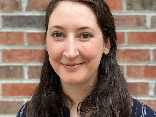 A headshot of a woman (Hannah Krieger) with long dark hair, smiling against a red brick wall background.