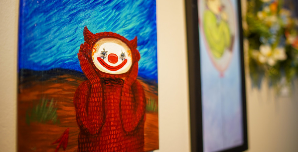 A small, square painting of a figure in a red, hooded devil costume with a tail, but the face is a clown face with a red nose and wide white smile. The figure is set against a dark, stormy blue and orange-brown background.