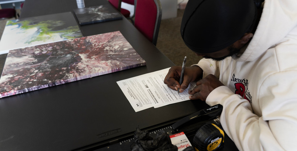 A high-angle shot focusing on an artist wearing a white hoodie and a black durag. He is carefully signing an official Arts Council "Public Works" submission form. Next to him on the table are two large abstract paintings with swirling pink, white, and green patterns.