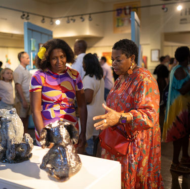 A lively scene inside the gallery during the exhibition. Two women—one in a vibrant floral dress and the other in a colorful geometric print—discuss a pair of sparkling, silver-toned ceramic sculptures (one of which is an octopus) displayed on a white pedestal.