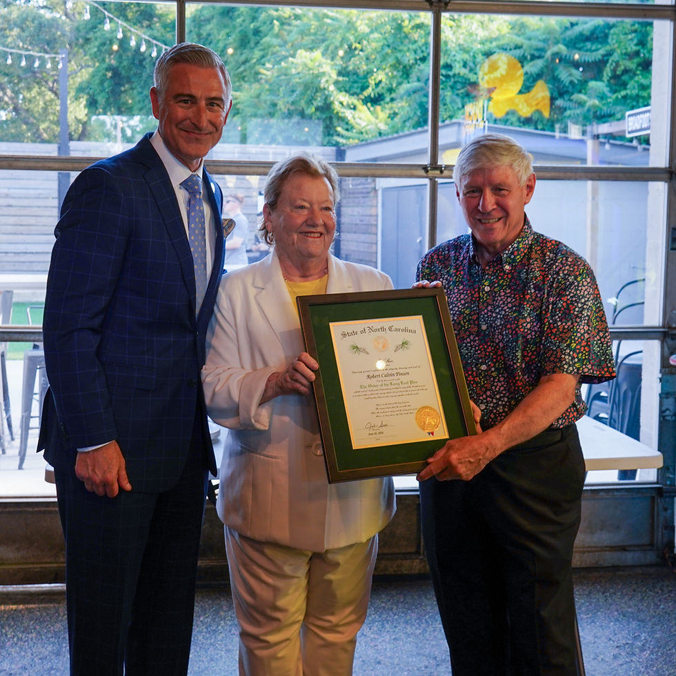 Three people smiling; two hold a framed certificate reading "State of North Carolina." Background shows trees and a window. Bright atmosphere.
