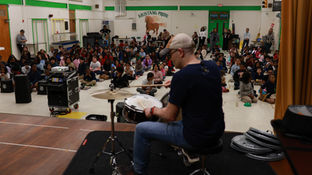 A drummer performs on stage as children sit watching in a school gym. The wall reads "Mustang Pride" with a horse mural. Mood is attentive.