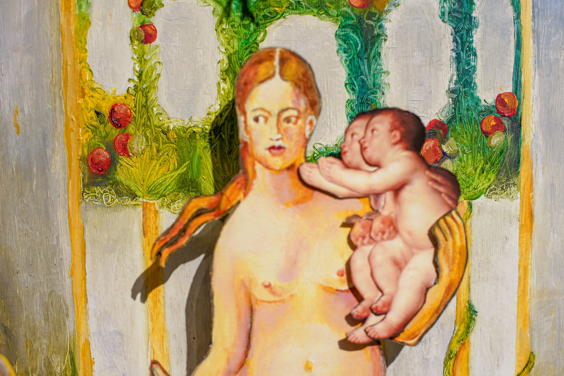 Close-up of the central figure in the diorama: a nude, fair-skinned woman with reddish-brown hair, holding two babies close to her chest. They are painted in front of a background of green vines and red fruit.