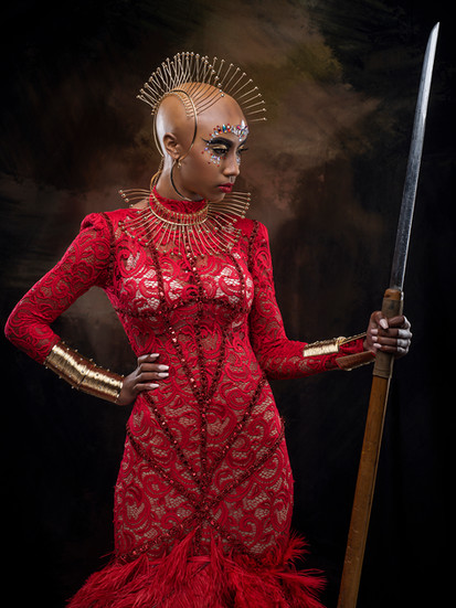 A dramatic portrait of a bald woman with elaborate metallic eye makeup and a gold wire headpiece, wearing a form-fitting, long-sleeved red lace gown with beading and a feathery skirt. She has gold cuffs on her forearms and a high, pointed, structured gold collar, and is holding a long, silver sword or spear against a dark, textured background