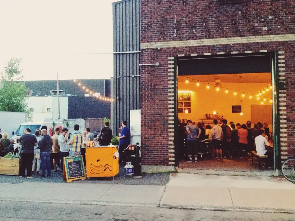People gather outside a brick building, some at a food stall. Inside, a crowd enjoys a well-lit space with string lights and menu boards.