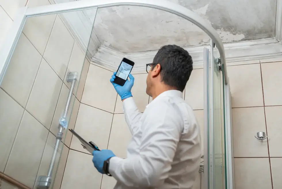 Gloved man in a tiled bathroom inspects a moldy ceiling with a phone and clipboard, focused and methodical.