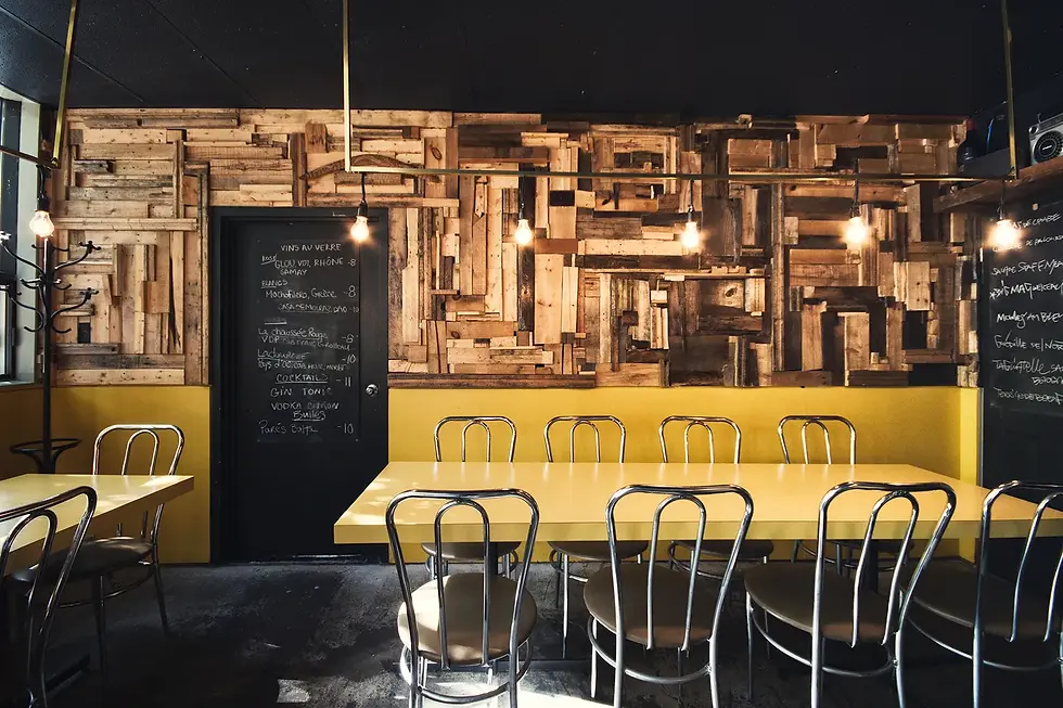 Restaurant interior with yellow tables, metal chairs, rustic wooden wall, hanging bulbs, and chalkboard menus. Cozy, vintage ambiance.