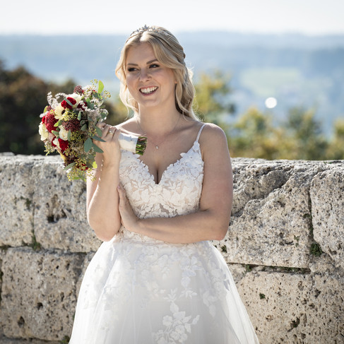 Braut steht vor Mauer, hält Brautstrauss hoch, lacht