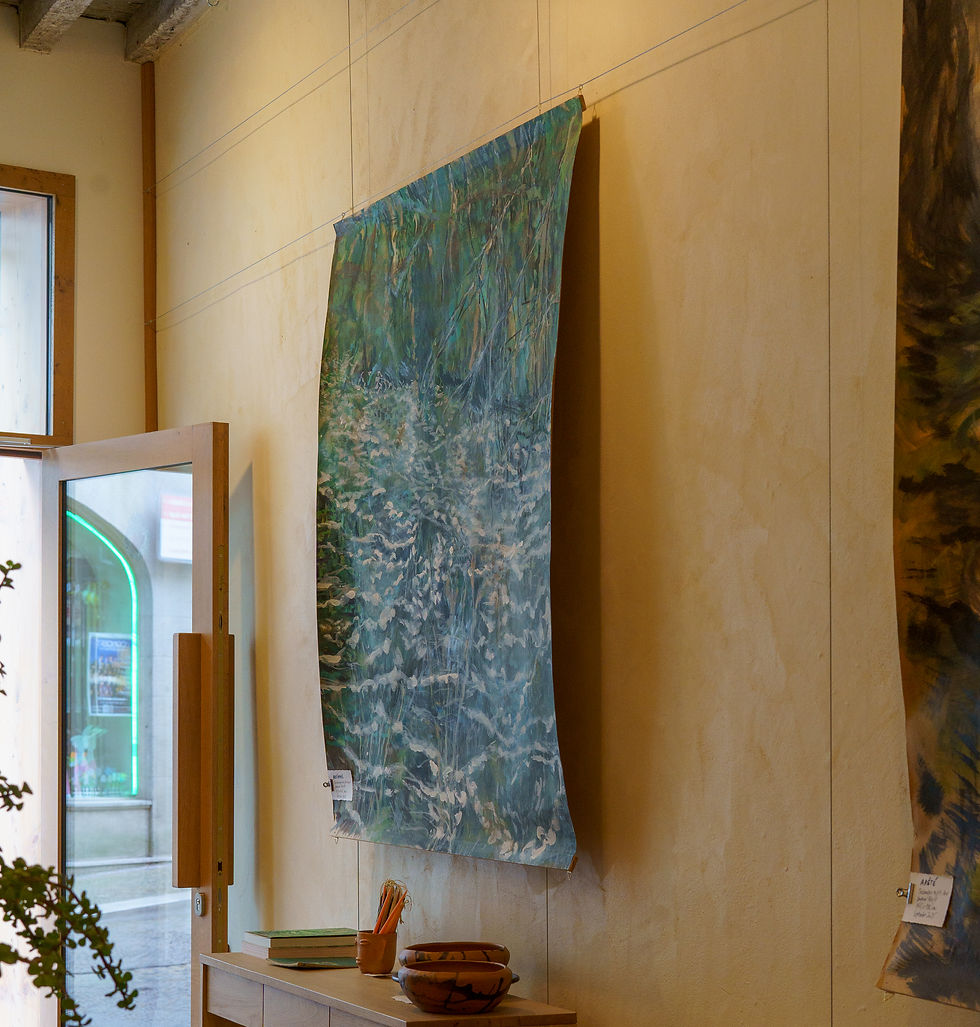 Tableau bleu-vert ondulé sur mur beige. Pot et livres posés sur une table en bois clair. Porte ouverte avec lumière extérieure. Galerie en plein coeur d'Arles.
