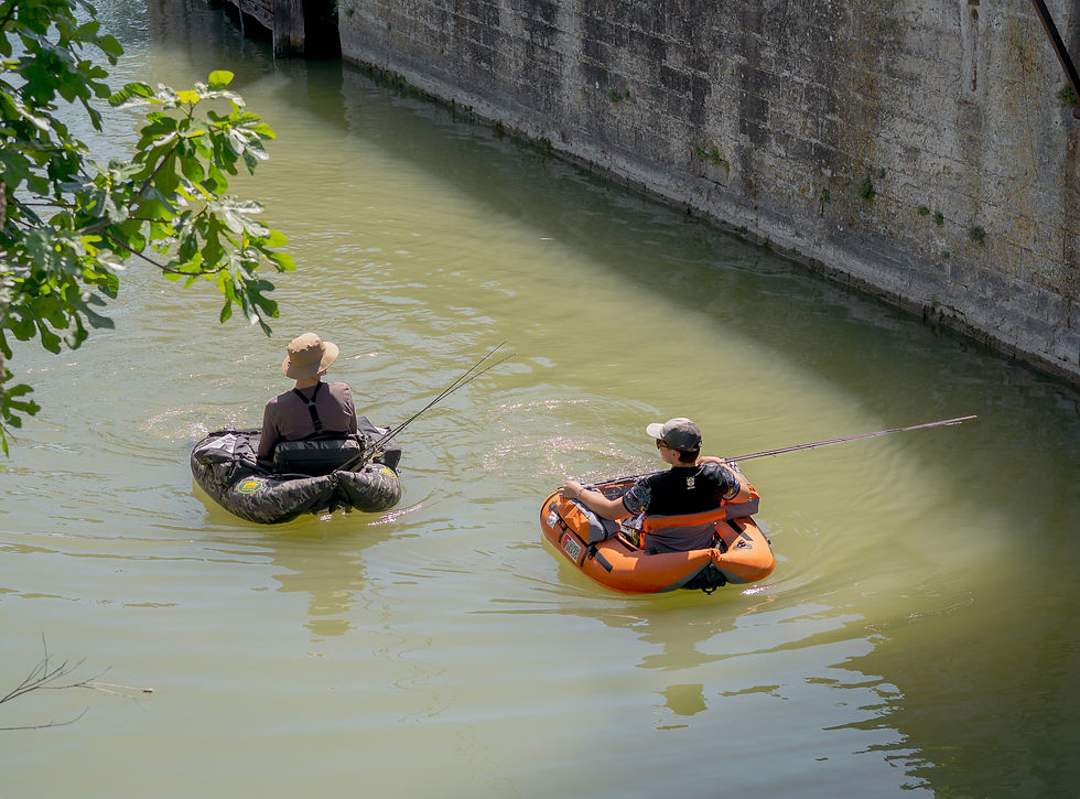 Deux personnes pêchent sur des bouées gonflables, l'une orange, l'autre vert foncé, dans un canal entouré de murs en pierre. Atmosphère paisible.