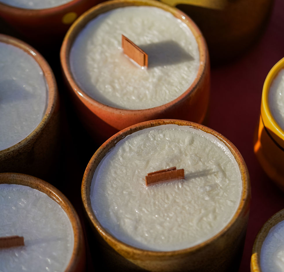 Bougies blanches dans des pots colorés, mèches en bois. Lumière naturelle, ambiance chaleureuse avec nuances de brun et orange.