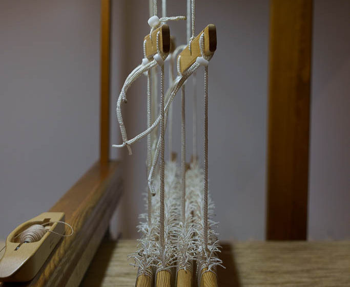 Métier à tisser en bois avec fils tendus blancs. Navette en bois sur la gauche. Ambiance artisanale dans une pièce éclairée.