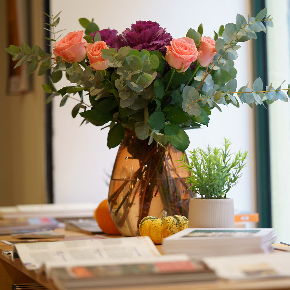 Bouquet de roses orange et de choux violets dans un vase brun, sur une table avec livres empilés, pot de plante et petites courges décoratives.