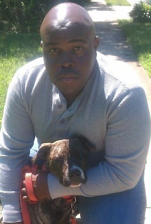 Carlton DeCosta kneeling on a sunlit sidewalk with his brindle-coated dog, Bernadette (Bernie), wrapped in a red harness."