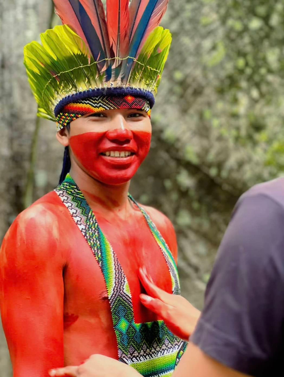 Un autochtone torse nue portant une peinture rouge sur le corps, avec une couronne de plume sur la tête et un collier artisanal en perle. Il y'a un homme qui pose sa main sur son torse. 