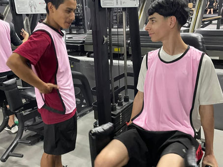 Dois jovens atletas de futebol conversando amigavelmente com coletes de treino durante rotina na academia do clube.