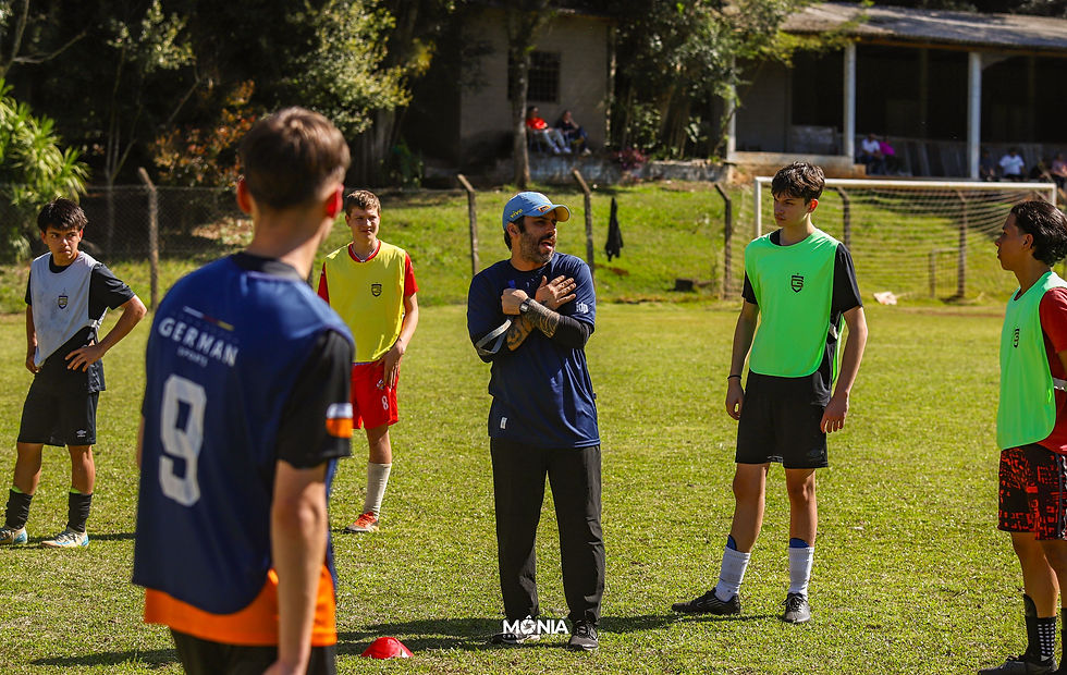 Treinador de futebol de boné azul e camiseta escura falando com grupo de atletas usando coletes coloridos no gramado durante avaliação.