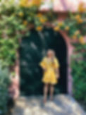 Woman in yellow dress standing before arched black door, flowers overhead.