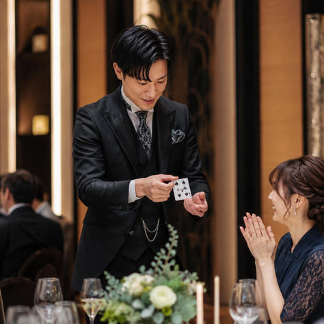 黒いスーツの男性マジシャンが宴会場でカード手品を披露し、向かいの女性が笑顔で拍手する温かな雰囲気。