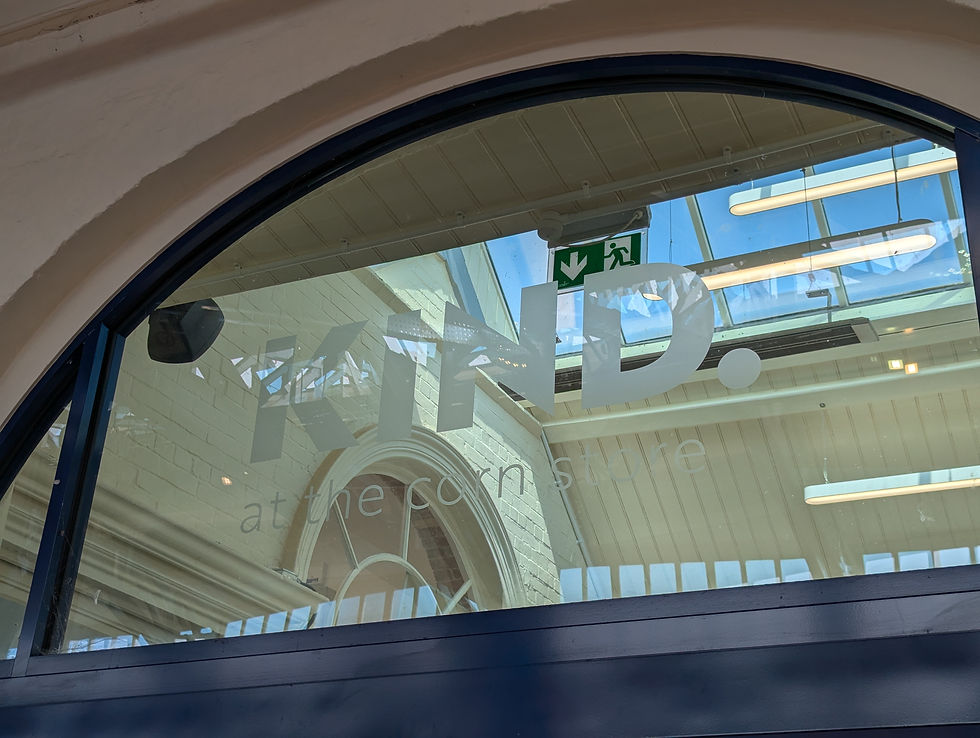 The entrance to Studio KIND. A glass arched window above the door displaying KIND at the Corn Store decal. Arched window with "KIND." text viewed from outside. Interior has a skylight and visible exit sign. Calm, daylight setting.