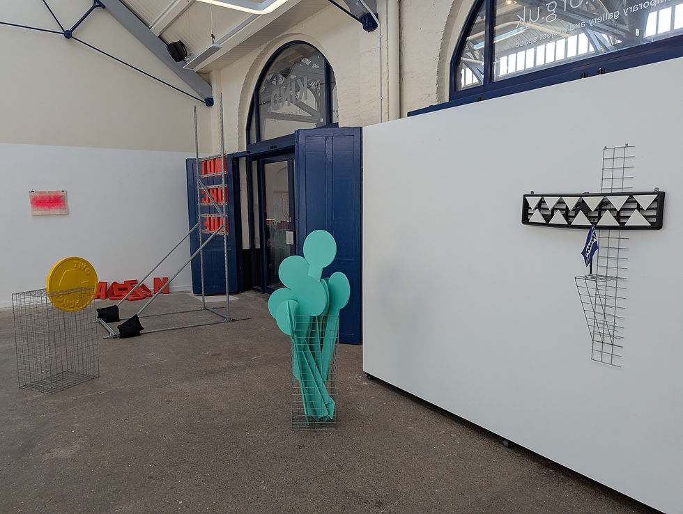 A shot of Stuar Robinson's exhibition. Art exhibit in a gallery with abstract sculptures: a yellow coin, aqua shapes, and geometric patterns. White walls, blue accents, and scaffolding.