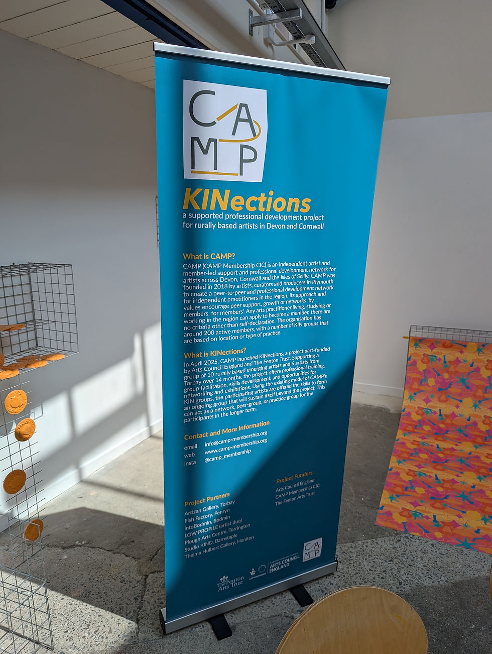 Blue banner for CAMP's KINections project detailing support for artists in Devon and Cornwall. Background includes art pieces and a chair.