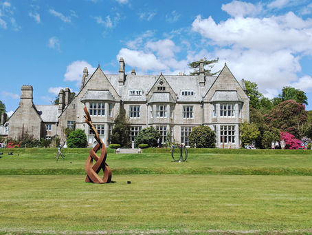A large stone mansion with manicured lawns, metal sculptures, and blooming bushes under a bright blue sky. People gather on the grass.