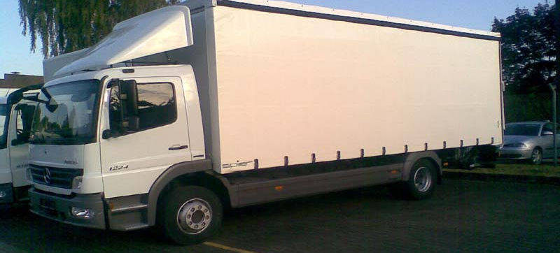 Weißer Lkw mit weißer LKW-Plane von Neumann Zelte und Planen, geparkt auf einem Parkplatz, im Hintergrund Bäume und weitere Fahrzeuge.