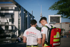 Ein Foto von hinten aufgenommen, das zwei junge männliche Sanitäter des SKALA Sanitätsdienstes zeigt, die an einem sonnigen Tag an der Spree-Promenade im Berliner Regierungsviertel stehen. Beide tragen weiße Poloshirts mit dem roten "SKALA PARAMEDICS SANITÄTSDIENST"-Logo und gelben Hosen. Der rechte Sanitäter trägt einen großen, roten PAX-Rettungsrucksack mit reflektierenden gelben Streifen. Sie stehen zusammen und blicken nach unten. Im Hintergrund links ist die moderne Architektur des Paul-Löbe-Hauses zu sehen, während der Berliner Fernsehturm und andere Gebäude die Skyline rechts im Hintergrund unter einem klaren blauen Himmel bilden. Ein Baumast ragt rechts oben ins Bild. Im Hintergrund am Ufer sitzt eine Person.