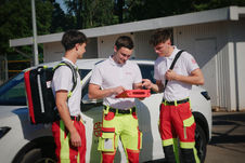 Drei junge Sanitäter in Dienstkleidung (weiße Poloshirts, rot-gelbe Einsatzhosen) stehen an einem weißen Einsatzfahrzeug und besprechen etwas auf einem Tablet in einer roten Schutzhülle. Der linke Sanitäter trägt einen großen Rettungsrucksack auf dem Rücken und blickt zu den anderen beiden. Der mittlere hält das Tablet, während der rechte Kollege mit einer Hand an seinen Rucksackgurt greift und auf den Bildschirm schaut. Das Logo auf ihren Shirts zeigt den Schriftzug „SKALA Paramedics“. Im Hintergrund ist ein weißes Gebäude im Sonnenschein zu sehen.