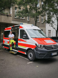 Ein lächelnder Rettungssanitäter steht mit verschränkten Armen in der geöffneten Schiebetür eines rot-weißen Volkswagen Rettungswagens von Skala Paramedics Sanitätsdienst; das Fahrzeug parkt am Straßenrand vor einem Wohngebäude.