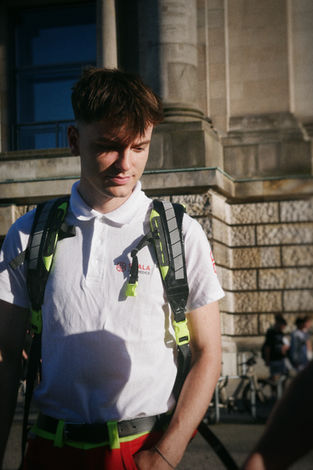 Ein junger Mann mit kurzen, braunen Haaren blickt nachdenklich nach unten. Er trägt die Uniform eines Rettungsdienstes, bestehend aus einem weißen Poloshirt mit einem roten Logo („SCALA PARAMEDICS“) auf der linken Brustseite und einer roten Hose mit neongrünen Gürtelschlaufen. Auf seinen Schultern trägt er die breiten, neongelb reflektierenden Gurte eines medizinischen Rucksacks. Das Bild ist im Hochformat aufgenommen; im Hintergrund ist die hellbraune Steinfassade eines historischen Gebäudes mit großen Säulen zu sehen. Das Sonnenlicht fällt von der Seite ein und wirft Schatten auf das Poloshirt.