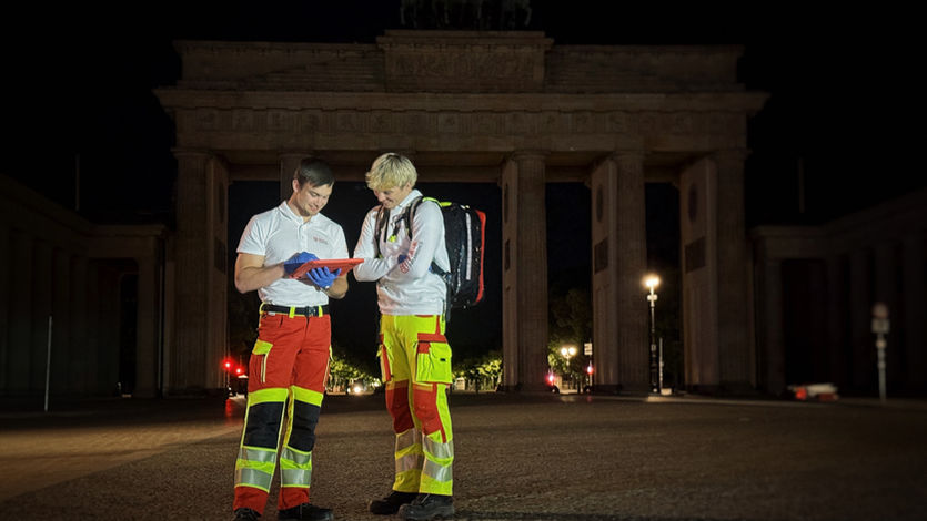 Nachtaufnahme in Berlin: Zwei männliche Einsatzkräfte des Sanitätsdienstes stehen auf dem Pariser Platz. Sie tragen weiße Oberteile und auffällige rot-gelbe Einsatzhosen mit Reflexstreifen. Das Brandenburger Tor ist im Hintergrund groß und dezent beleuchtet zu sehen. Die Sanitäter nutzen ein Tablet zur Dokumentation oder Koordination, einer trägt zudem einen großen schwarzen Rettungsrucksack.
