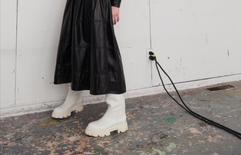 A model posing in a long black leather dress and chunky white boots from the Tanya Taylor Fall 2022 collection