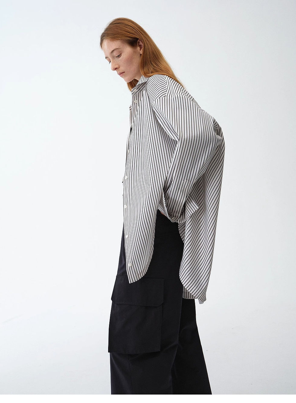 Thumbnail: Model in side profile wearing  asymmetric striped button-down shirt with black trousers