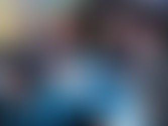 A imagem mostra quatro homens sentados ao redor de uma mesa azul em um ambiente de evento, sorrindo para a câmera. À esquerda, um deles veste camiseta preta com a logo da Logcomex e crachá no pescoço; ao lado, um homem de terno azul-marinho; ao centro, outro homem de terno preto fazendo sinal de positivo; e à direita, um homem de terno cinza. Sobre a mesa, há papéis, post-its coloridos e um celular. Ao fundo, diversas outras mesas com pessoas em conversas e interações, sugerindo um espaço de networking ou rodada de negócios. O ambiente é iluminado por luzes suaves, com projeções nas paredes e um clima dinâmico e colaborativo