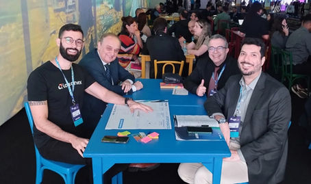 A imagem mostra quatro homens sentados ao redor de uma mesa azul em um ambiente de evento, sorrindo para a câmera. À esquerda, um deles veste camiseta preta com a logo da Logcomex e crachá no pescoço; ao lado, um homem de terno azul-marinho; ao centro, outro homem de terno preto fazendo sinal de positivo; e à direita, um homem de terno cinza. Sobre a mesa, há papéis, post-its coloridos e um celular. Ao fundo, diversas outras mesas com pessoas em conversas e interações, sugerindo um espaço de networking ou rodada de negócios. O ambiente é iluminado por luzes suaves, com projeções nas paredes e um clima dinâmico e colaborativo