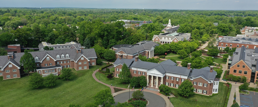 Aerial view of a TCNJ campus with brick buildings, lawns, and Trenton Hall at center under a cloudy sky.