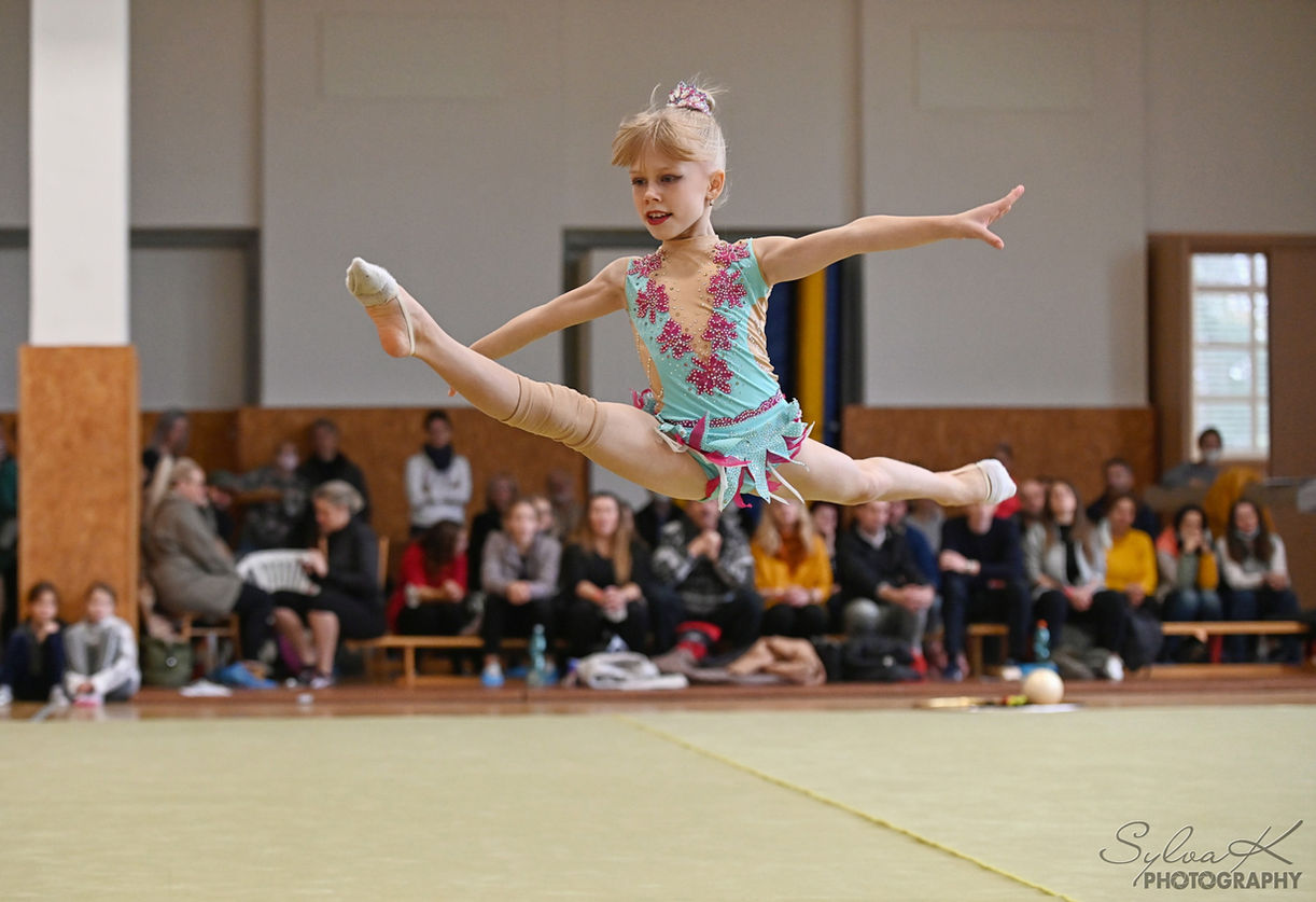Dětský sport, reportáž z pohárové soutěže. Gymnastka během sestavy bez náčiní, zachycená ve skoku. Blondýnka v tyrkysovém gymnastickém dresu. Foto Sylva Křenková, Olomouc.