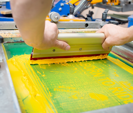 Hands using a squeegee to apply yellow ink through a screen stencil during a screen printing process, with the words "BORN TO