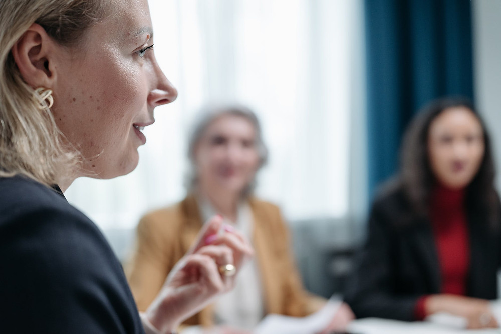 Three women in a meeting, two of them being blurred on the background, holding white papers, with brown and white and black and red outfits and a focused white blonde women on the right side of the picture talking with her left hand slightly raised