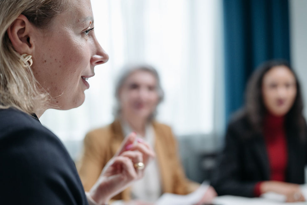 Three women in a meeting, two of them being blurred on the background, holding white papers, with brown and white and black and red outfits and a focused white blonde women on the right side of the picture talking with her left hand slightly raised