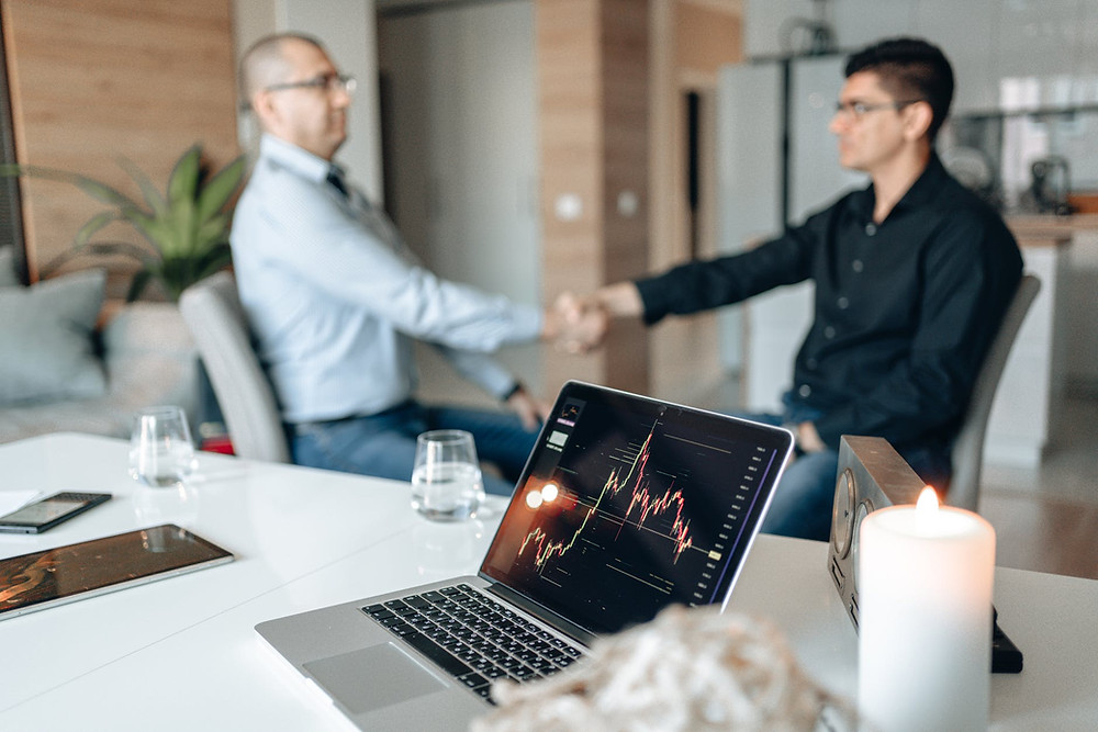 Office setting with a computer, showcasing a graphic, open on a white table ando two sitting men, shaking hands, on the back, unfocused