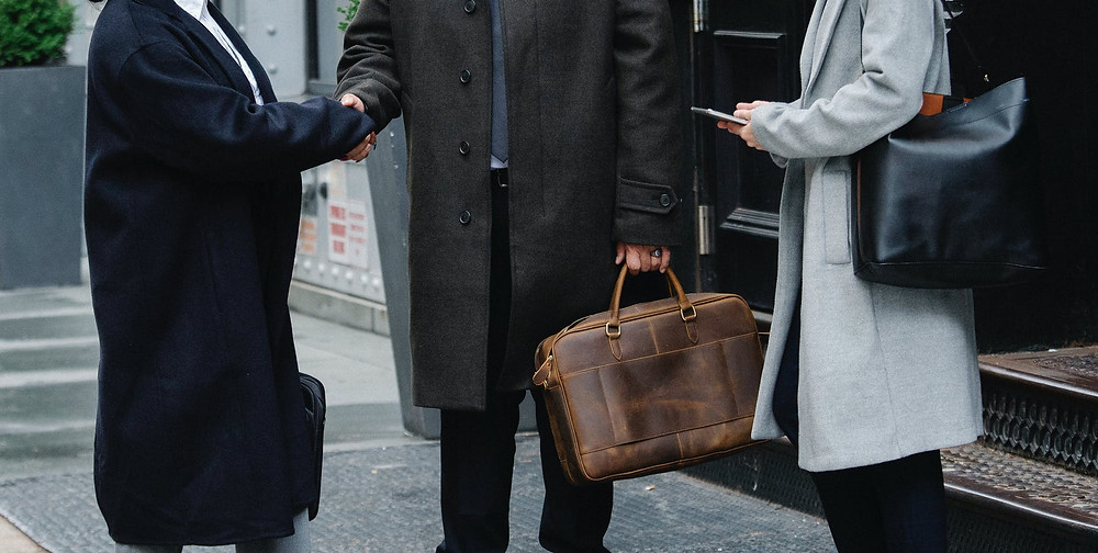 two men, both wearing black coats shaki, one of them holding a brown bag and shaking hands and a woman on the right with a black bag on her left side and a gray coat
